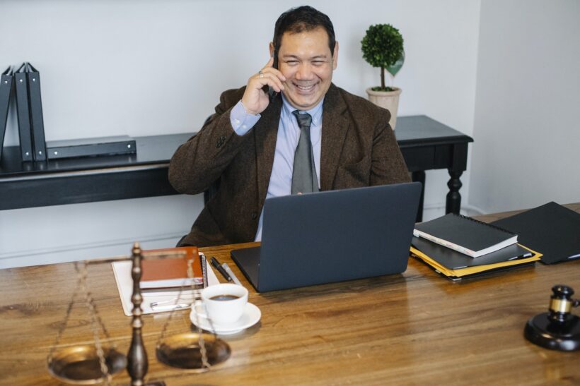 Joyful Asian male lawyer wearing formal suit having conversation on mobile phone and using laptop while working in modern law firm
